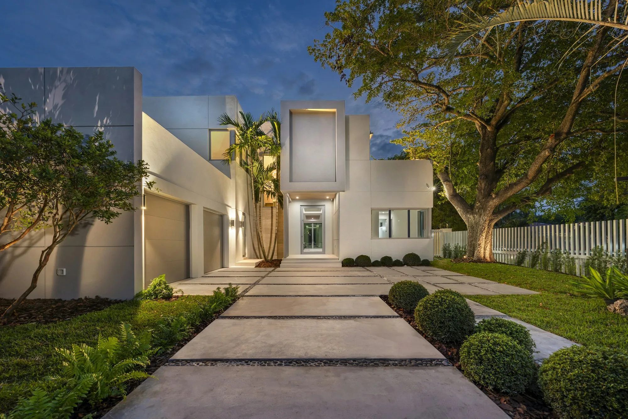 Lighted walkway leading to a modern white house with palms